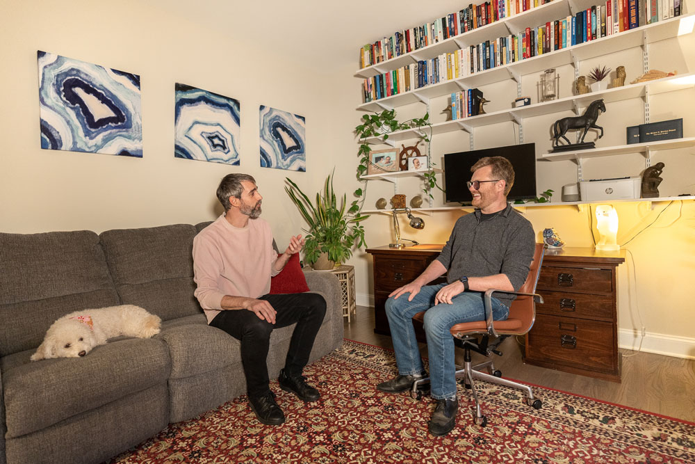 Photo of two men sitting in a home office talking. Next to one, a sleeping dog lies on the couch.