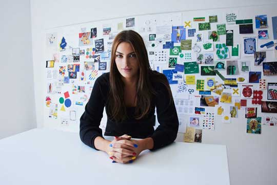 Photo of a woman dressed in black sitting at a table, staring directly at the camera with hands folded and a slight smile.