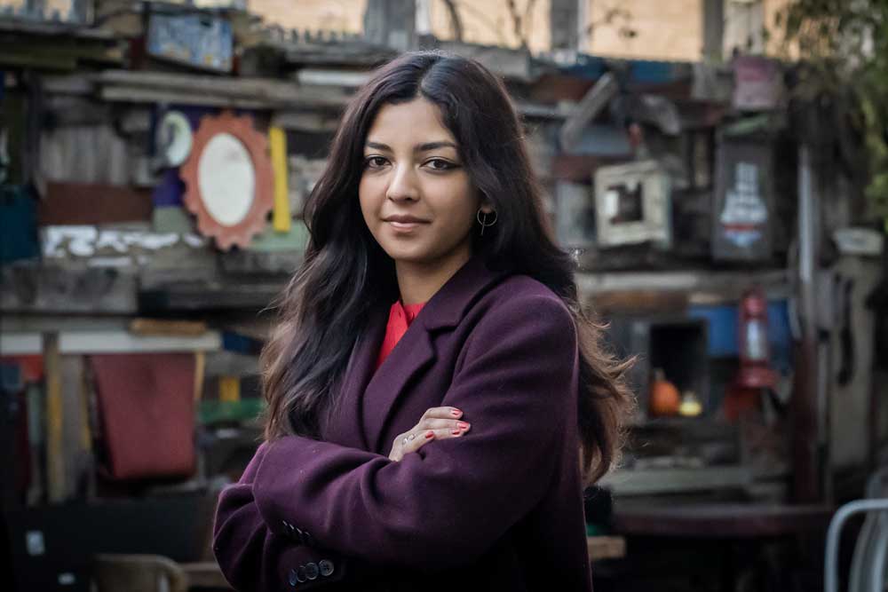 Photo of a woman dressed in a purple peacoat with a red undershirt, staring at the camera with arms crossed and a slight smile.