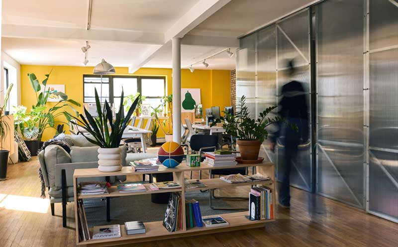 Photo of a sunlit office with a bright, yellow wall in the background. A blurred figure is passing through the space on the right.