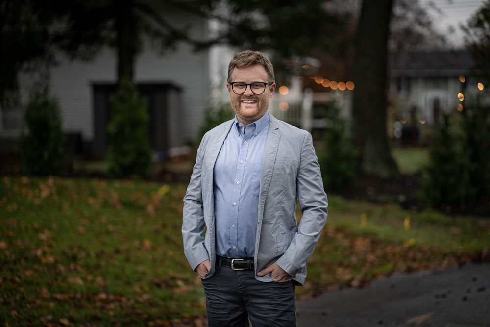 Photo of a man with glasses dressed in a gray blazer and dark gray pants standing in a yard, hands in pockets, with a wide smile.